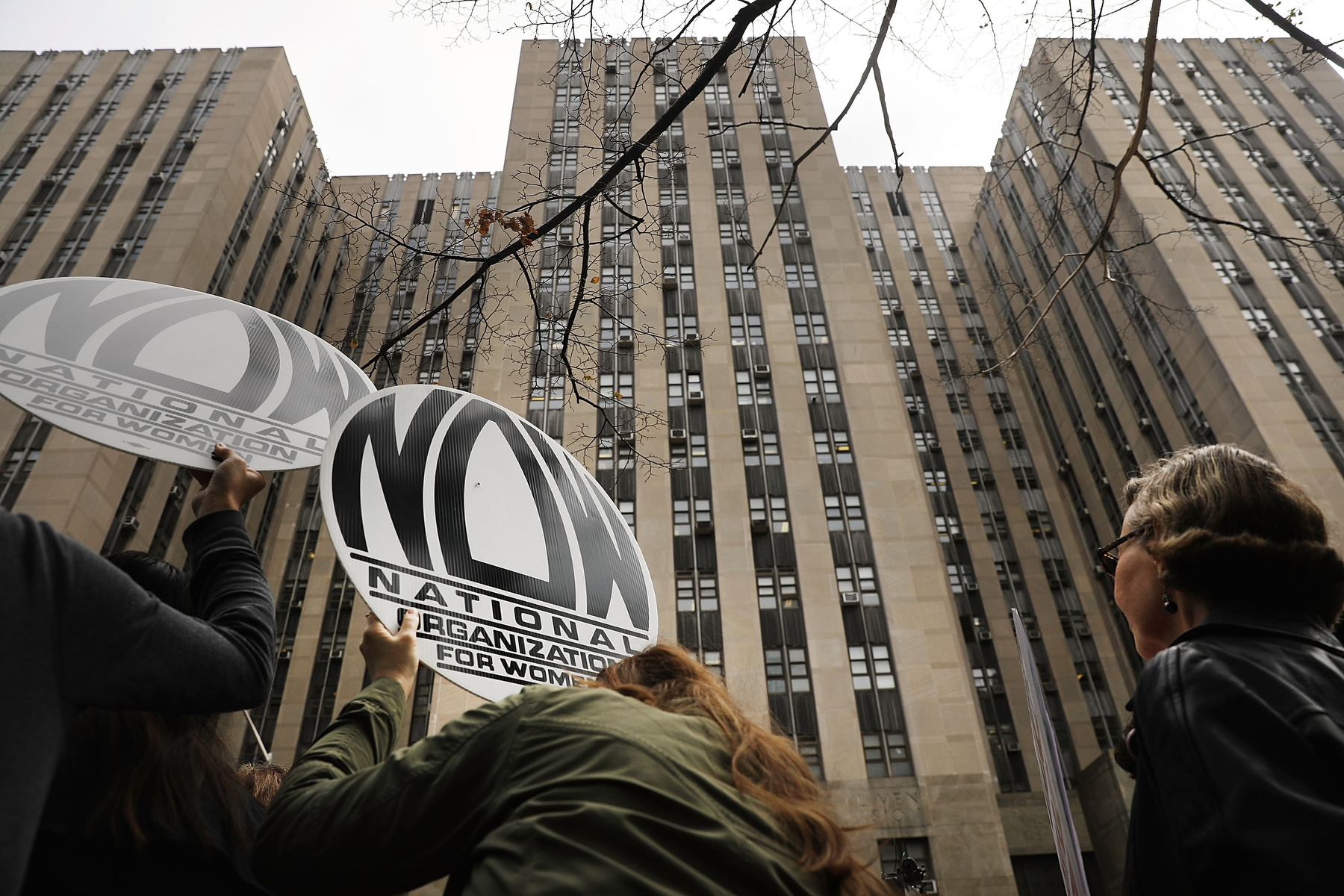 Women hold signs saying "NOW" in front of a building.