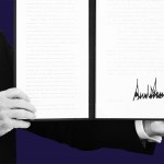 A close-up of President Trump holding open a signed executive order with a large signature visible on the right page.