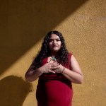 Angélica Reyes stands with her hands over her chest in front of a textured yellow wall.