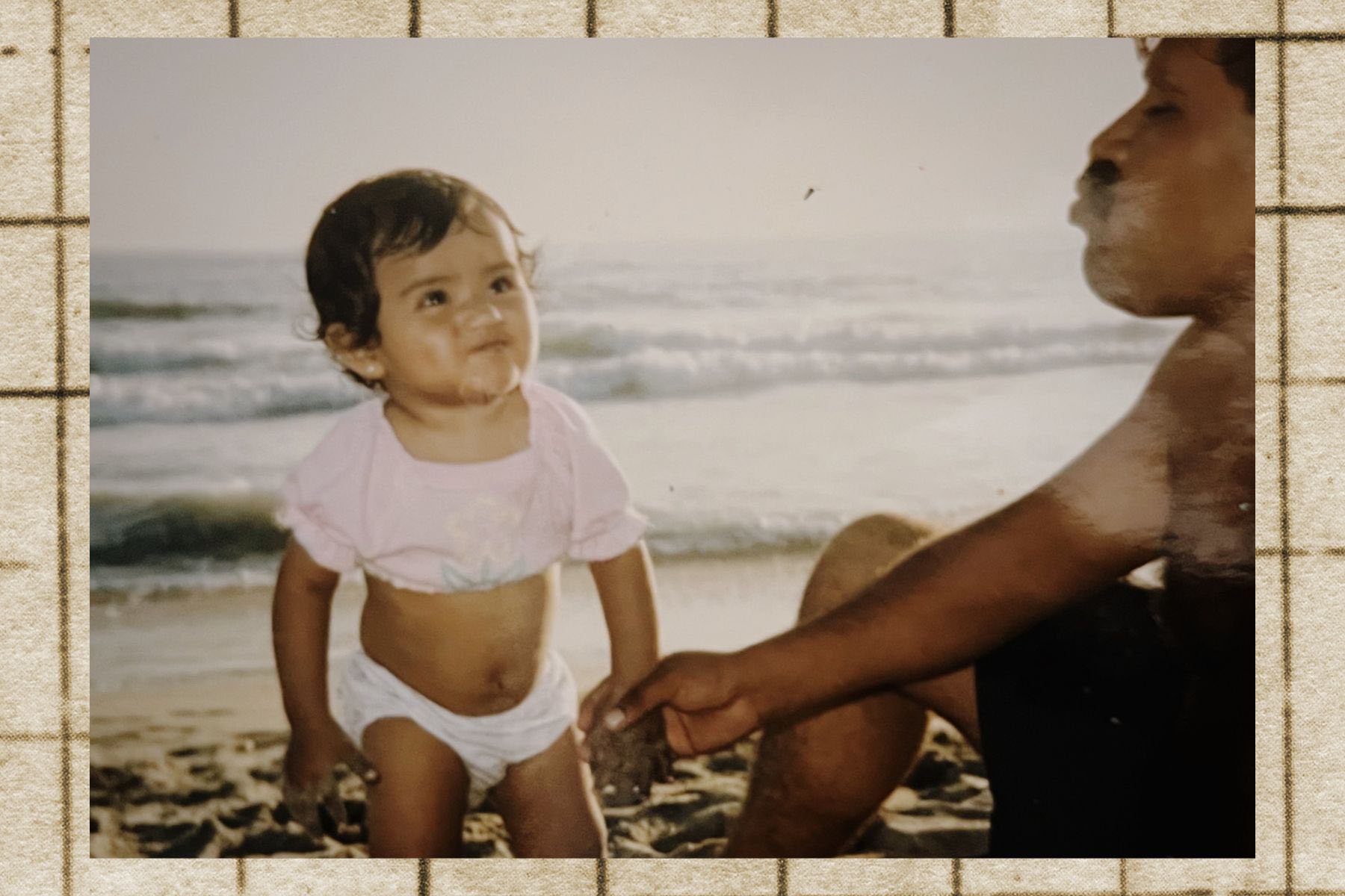 A toddler-aged Reyes stands in the sand at the beach, smiling toward her father, who is partially visible and holding her hand.