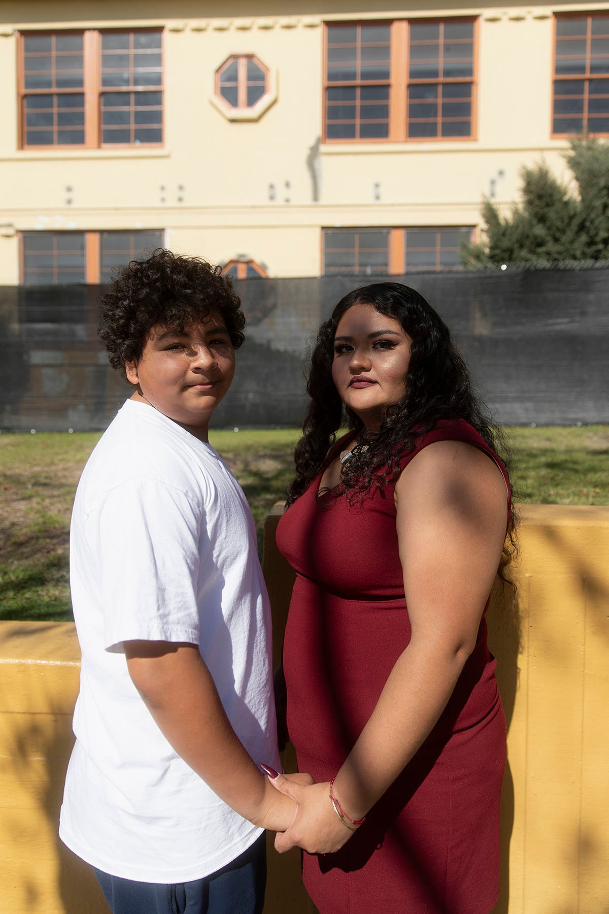 Reyes and her teenage son Nathan stand side-by-side holding hands in front of a yellow school building, both looking directly at the camera.