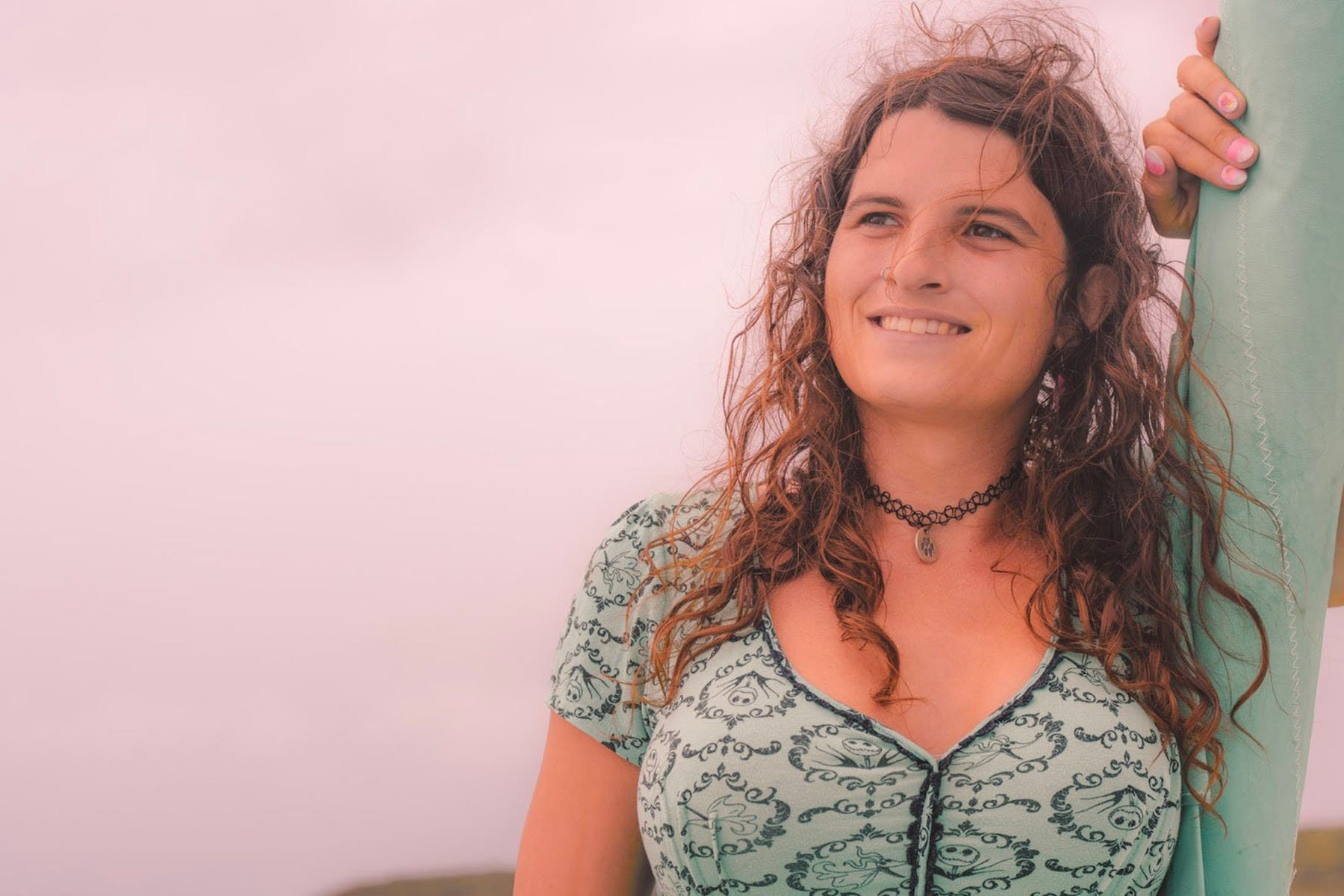 A close-up of a smiling Kelsey Granger with curly hair, wearing a mint green patterned dress and a black choker necklace.