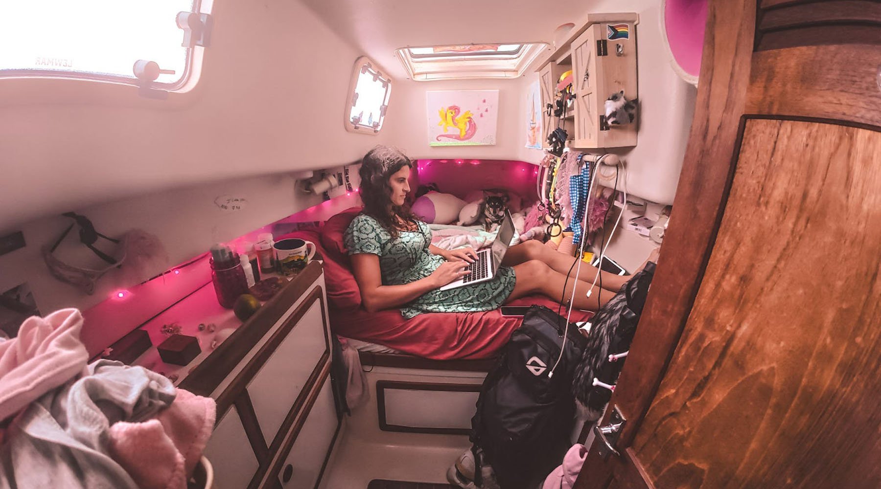 Kelsey Granger sits on a bed in a small, cozy boat cabin with pink lights and decorations, typing on a laptop with a dog lying next to her.
