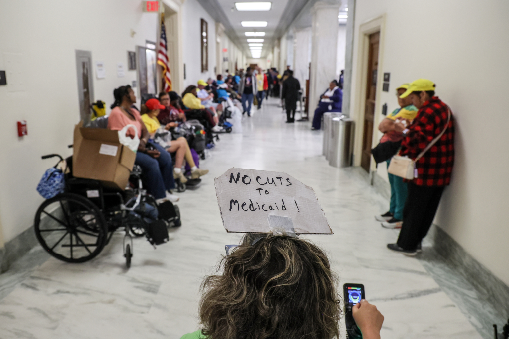 A person using a wheelchair wears a "no cuts other than Medicaid" placard on their head.
