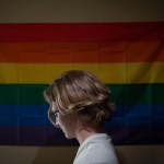 A young trans person in front of a rainbow flag.