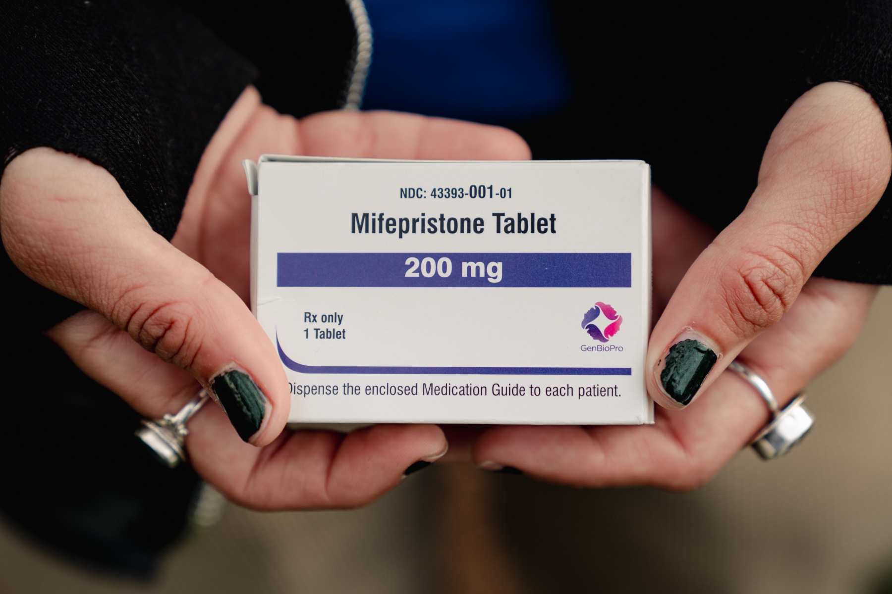 Hands with green nail polish wearing silver rings holding a box containing a tablet of mifepristone.