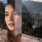 Jewel Lazaro, a woman with long dark hair, looks out a window her face partially reflected in the glass, with mountains and a balcony visible in the background.