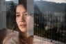 Jewel Lazaro, a woman with long dark hair, looks out a window her face partially reflected in the glass, with mountains and a balcony visible in the background.