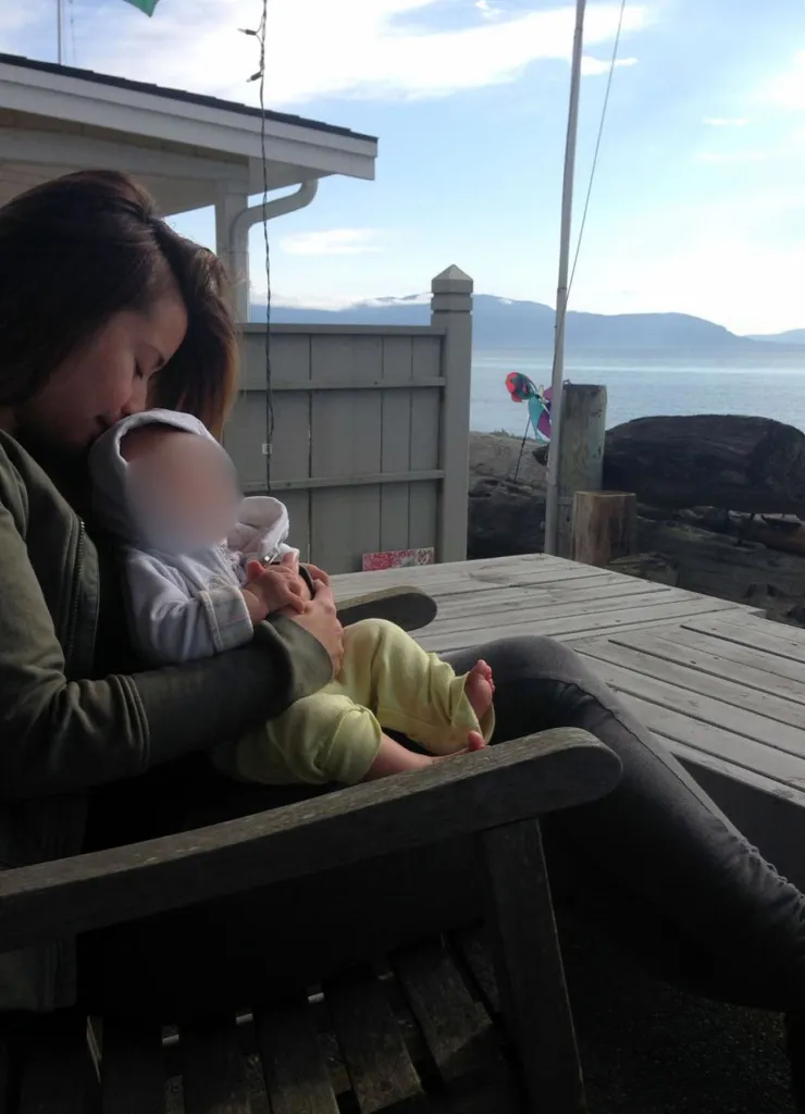 Jewel Lazaro sits outdoors on a wooden bench, cradling and kissing Lucia, whose face is blurred; behind them is a waterfront view with mountains.