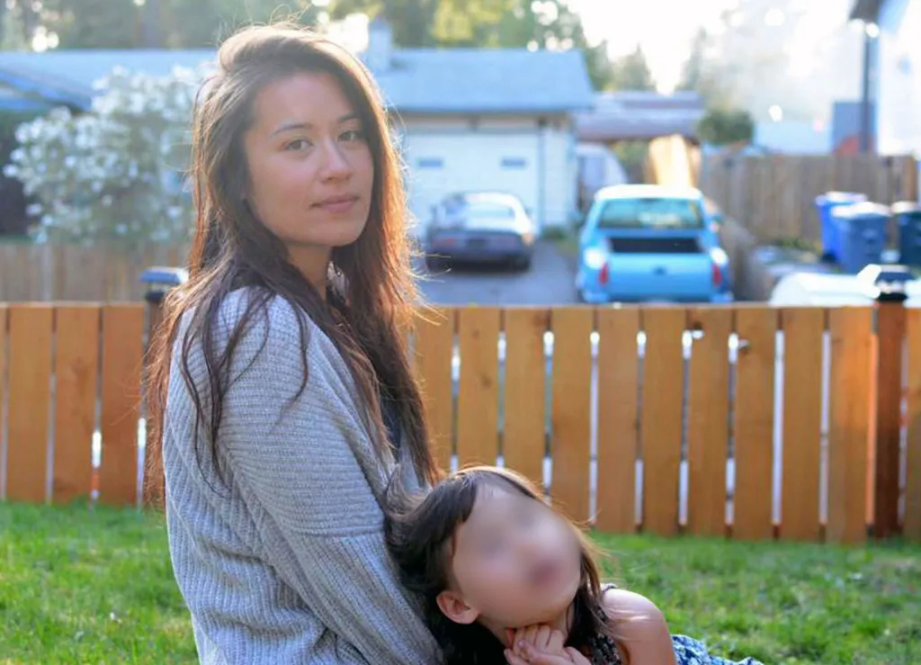 Jewel Lazaro sits on grass next to a wooden fence, her arm around Lucia, whose face is blurred and who leans against her.