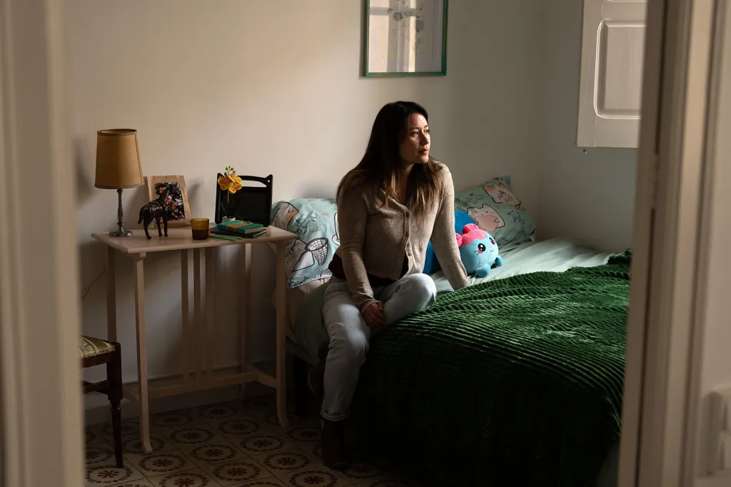 Jewel Lazaro sits on a bed in a room decorated with stuffed animals and colorful bedding, looking out toward the window.