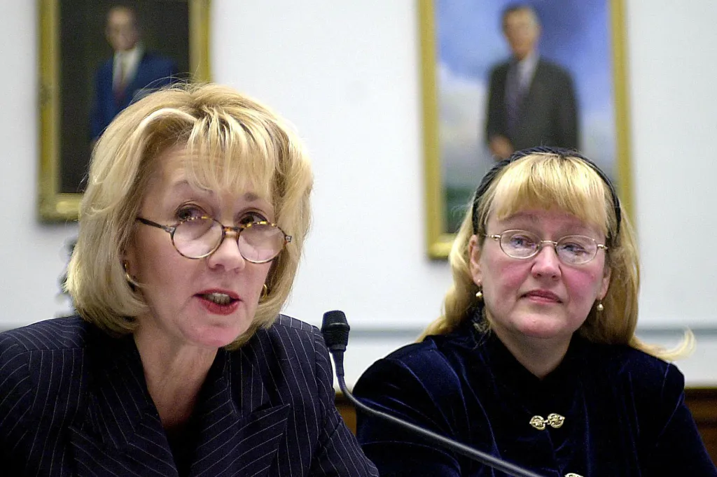 Two women sit at a congressional hearing; one speaks into a microphone while the other listens, both wear glasses and formal attire.