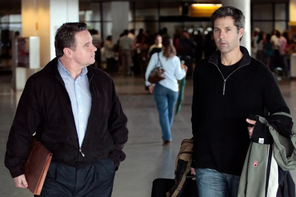 David Goldman, wearing a black sweater, walks through a busy airport terminal alongside Rep. Chris Smith, who is holding a briefcase and looking at Goldman. Travelers are visible in the background.