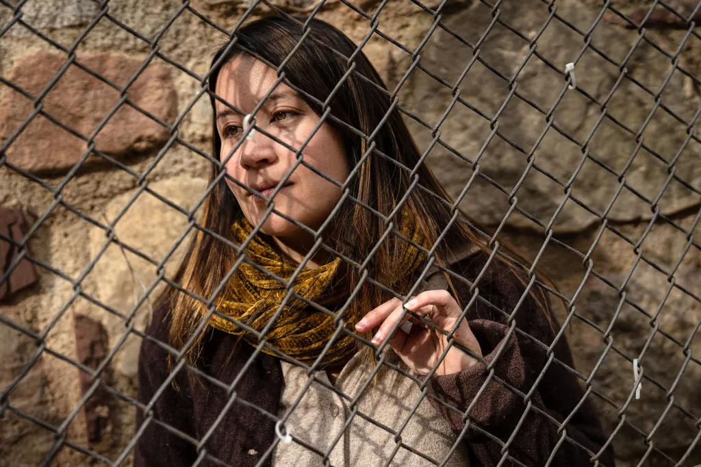 Jewel Lazaro gazes thoughtfully through a chain-link fence at a park near her apartment in Spain. Shadows of the fence fall across her face.