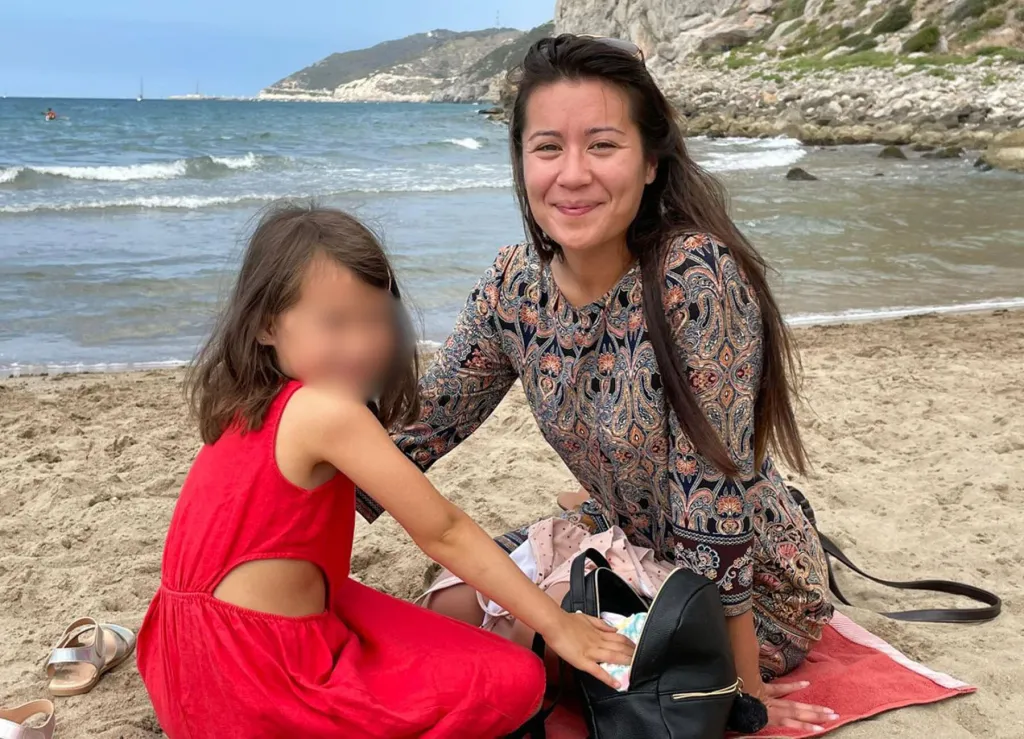 Jewel Lazaro smiles while sitting on a towel at a beach in Spain, next to her daughter Lucia, who is wearing a red dress and has her face blurred.