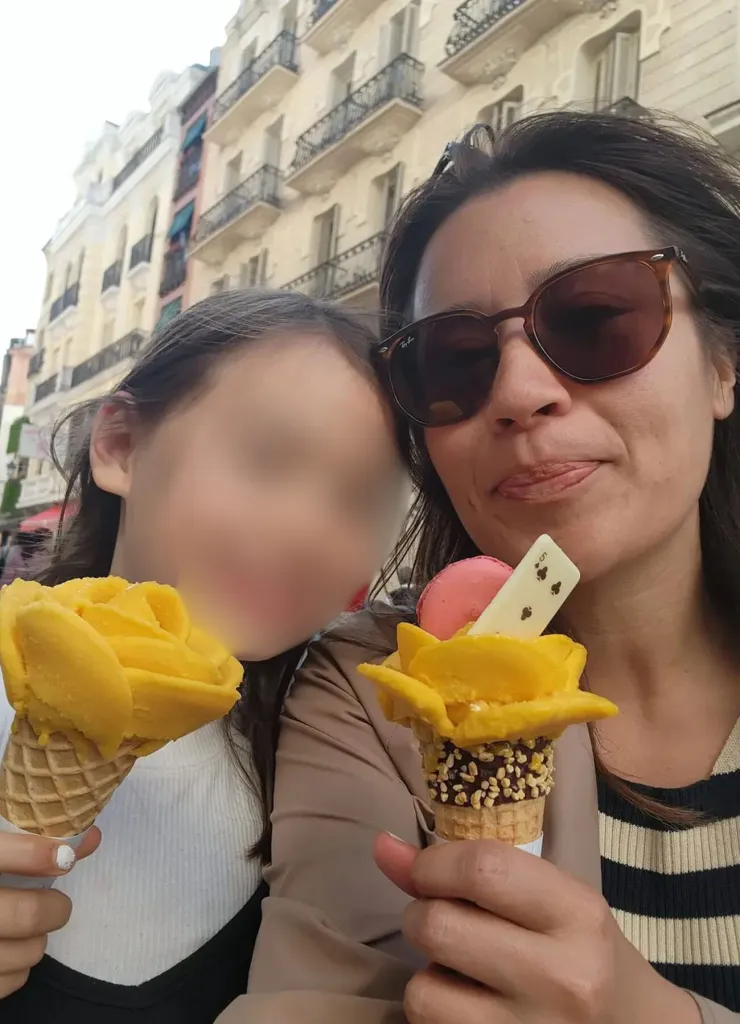 Jewel and Lucia enjoy ice cream shaped like yellow flowers in waffle cones as they pose for a selfie on a city street in Madrid. Lucia’s face is blurred.