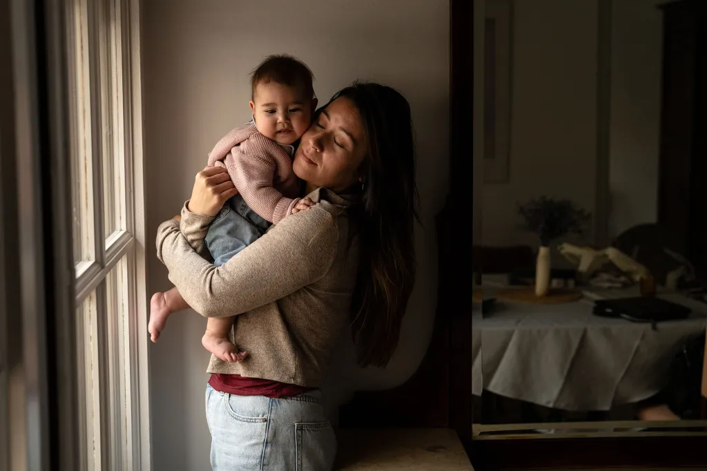 Jewel Lazaro holds her baby Selah close near a window in their apartment. Jewel’s eyes are closed as she hugs Selah, who looks out with a calm expression.