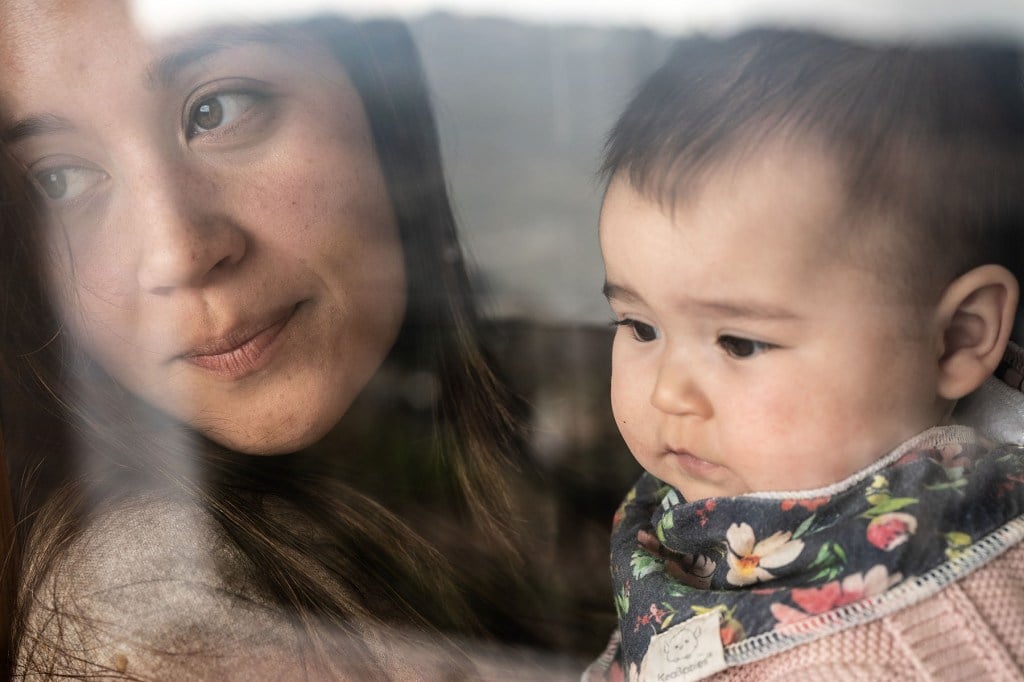 Jewel Lazaro and baby Selah look out a window together. Jewel’s face is partially reflected in the glass as she glances down, while Selah gazes outward.