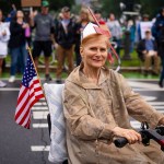 An older woman in a rain poncho rides a mobility scooter, smiling and holding an American flag during a protest. People with signs and flags are visible in the blurred background.