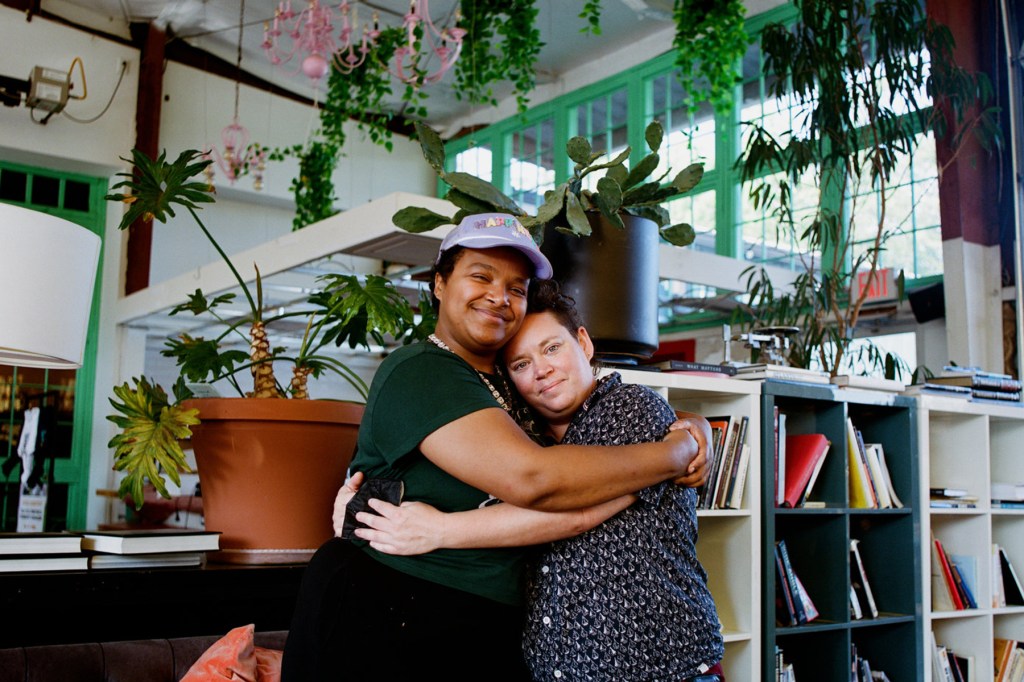 Two people hug each other against a backdrop of books and plants.