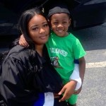 A woman in a black graduation gown kneels and smiles softly at the camera while hugging a young boy.