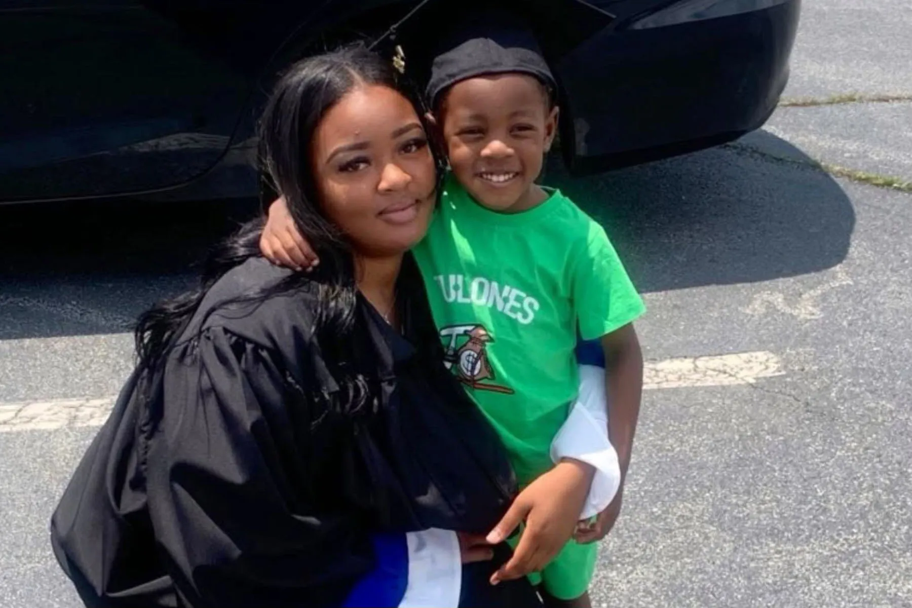 A woman in a black graduation gown kneels and smiles softly at the camera while hugging a young boy.