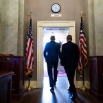 Two politicians walk through a door, seen from behind with a clock above the door.
