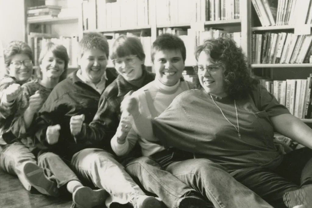 A row of people lean into each other while seating on the floor in front of stacks of books.