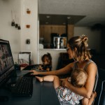 A woman holds a child while working at the computer as another child sits next to her.