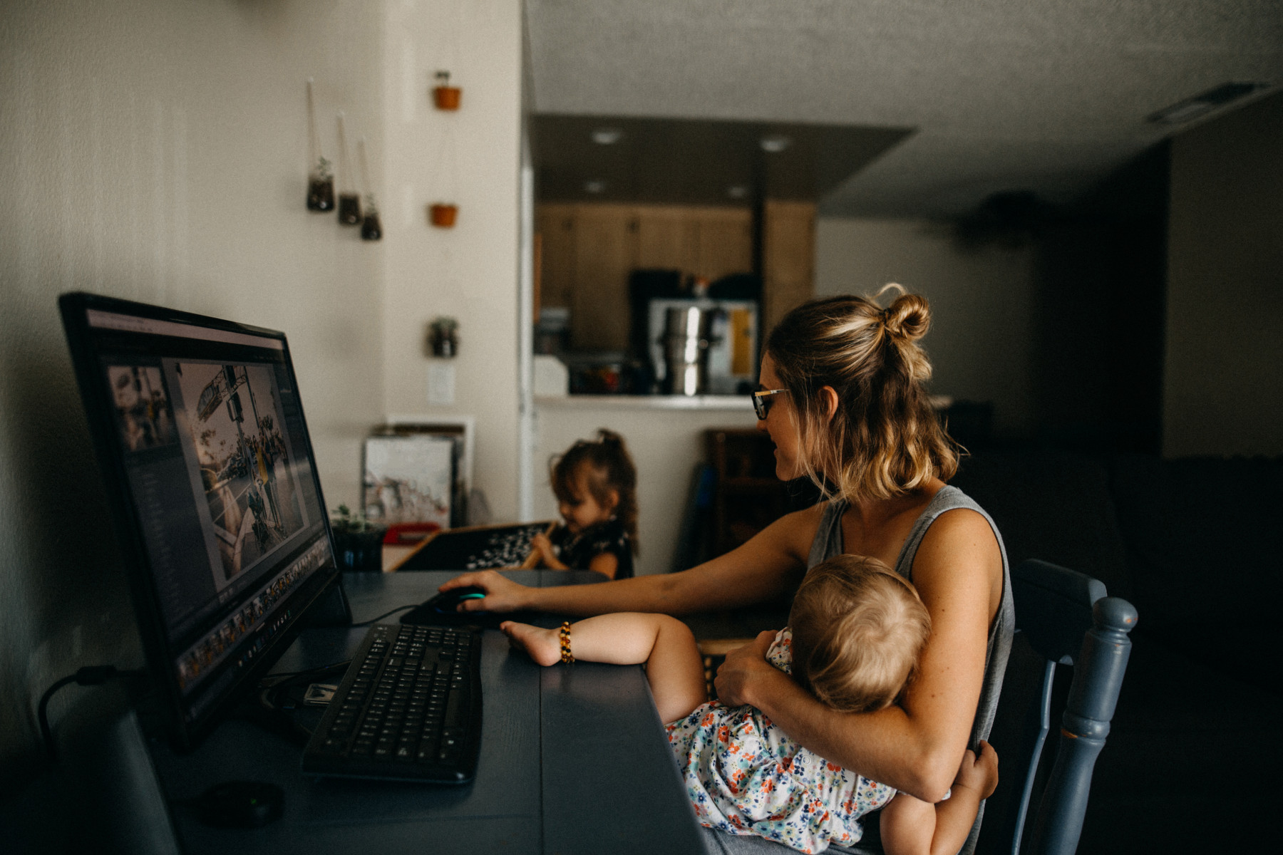 A woman holds a child while working at the computer as another child sits next to her.