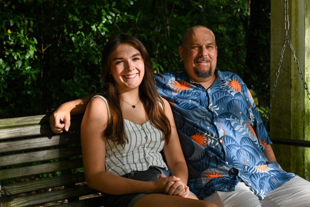 A daughter and her dad sit on a porch swing, the father's arm around his daughter's shoulders as they both smile.