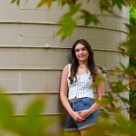 A girl leans against a concrete structure, framed by leaves. She is wearing a white striped tank top and denim shorts.