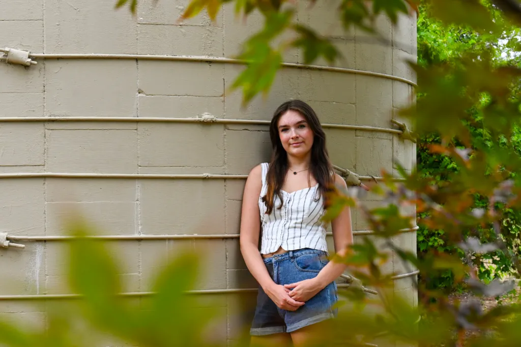 A girl leans against a concrete structure, framed by leaves. She is wearing a white striped tank top and denim shorts.