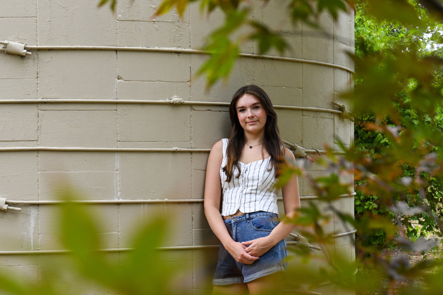 A girl leans against a concrete structure, framed by leaves. She is wearing a white striped tank top and denim shorts.