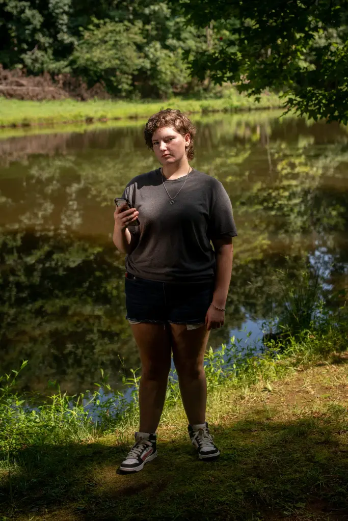 A person stands holding a phone by a pond.