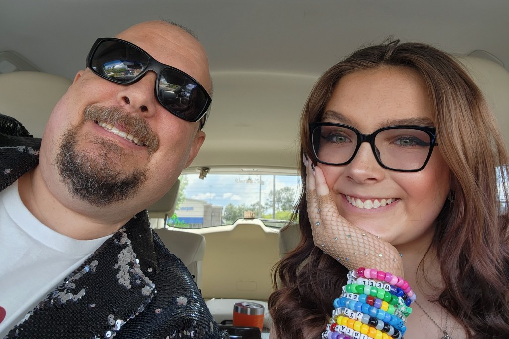 A man wearing sunglasses takes a selfie with his daughter wearing black eyeglasses, a sparkly glove and resting her shin on her hand and a wrist full of beaded bracelets.