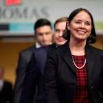 Shenna Bellows smiles, walking down a hallway surrounded by men in suits and military uniforms.