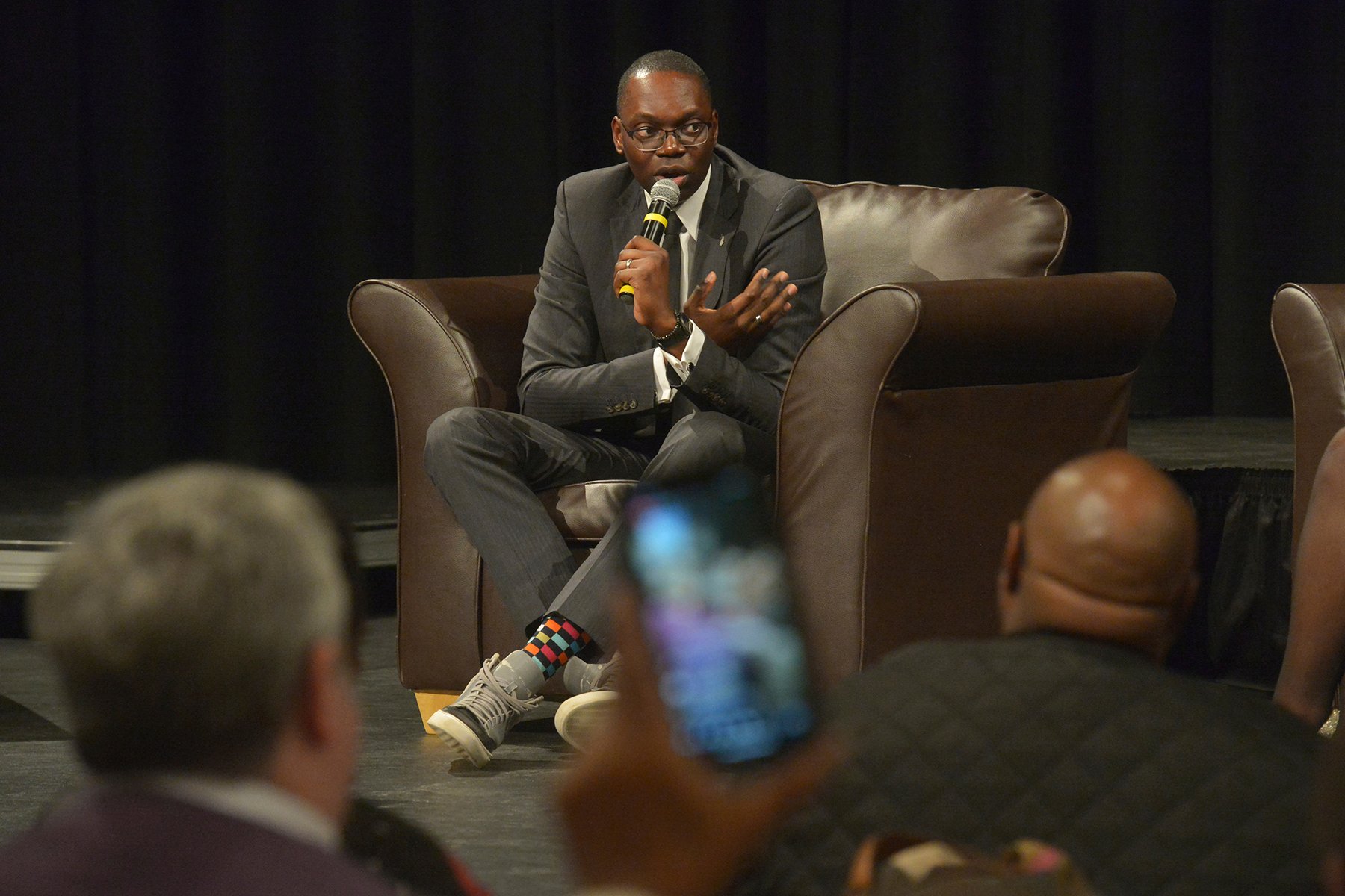 Garlin Gilchrist sits onstage in a suit and sneakers, speaking to a crowd.