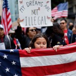 A young girl holds a large American flag during a pro-immigration rally, with a crowd of demonstrators behind her holding U.S. flags and signs. One prominent sign in the background reads “MY DREAMS ARE NOT ILLEGAL.”