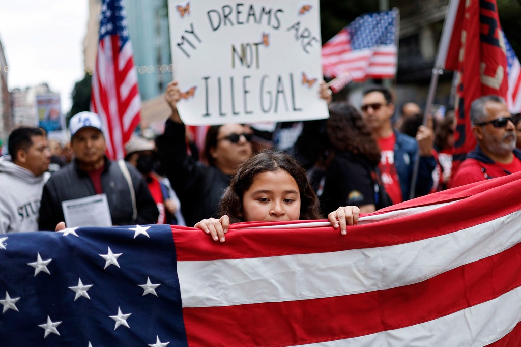 A young girl holds a large American flag during a pro-immigration rally, with a crowd of demonstrators behind her holding U.S. flags and signs. One prominent sign in the background reads “MY DREAMS ARE NOT ILLEGAL.”