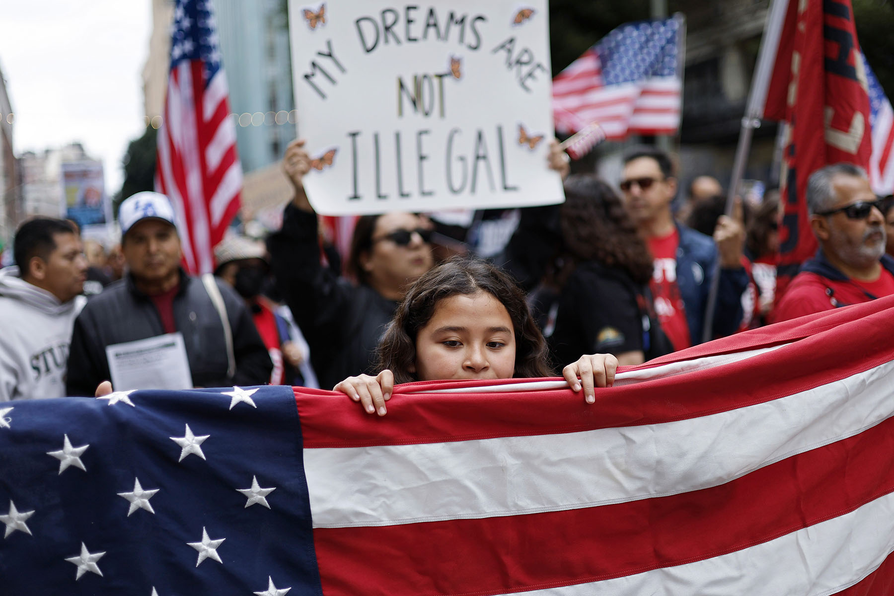 A young girl holds a large American flag during a pro-immigration rally, with a crowd of demonstrators behind her holding U.S. flags and signs. One prominent sign in the background reads “MY DREAMS ARE NOT ILLEGAL.”