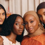 Four black and brown women pose closely together, looking directly at the camera. They have glowing skin and wear earth-toned clothing and gold jewelry. Two wear hijabs.