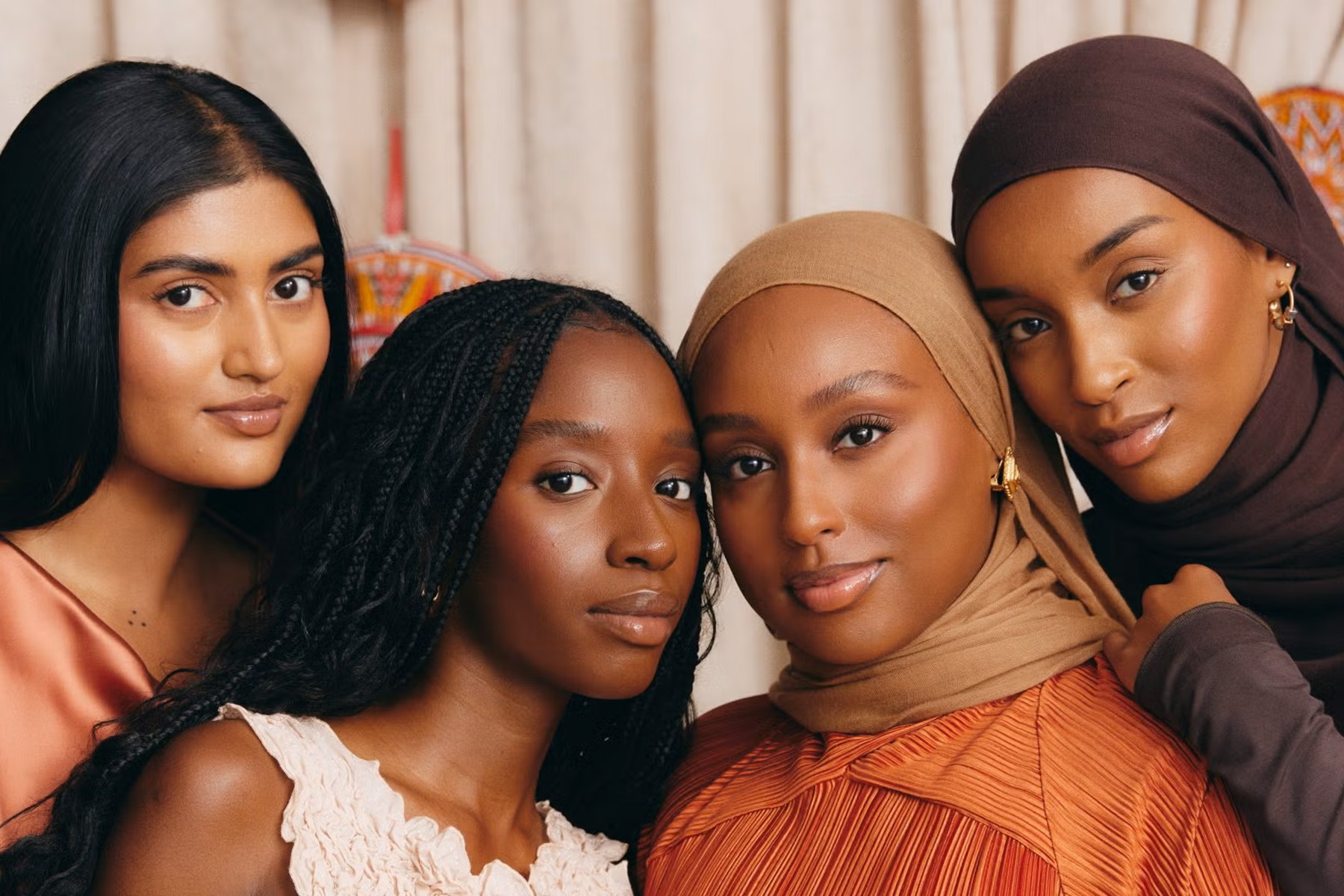 Four black and brown women pose closely together, looking directly at the camera. They have glowing skin and wear earth-toned clothing and gold jewelry. Two wear hijabs.