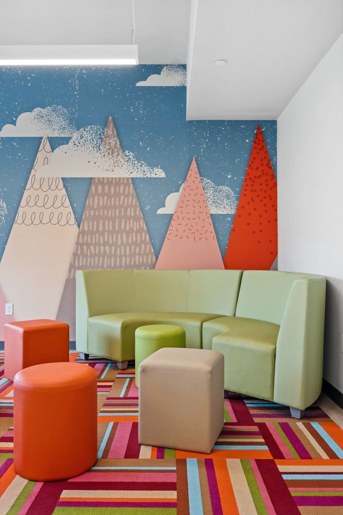 A colorful seating area with a patterned rug, pastel furniture, and a mural of trees and clouds inside the children’s lounge.
