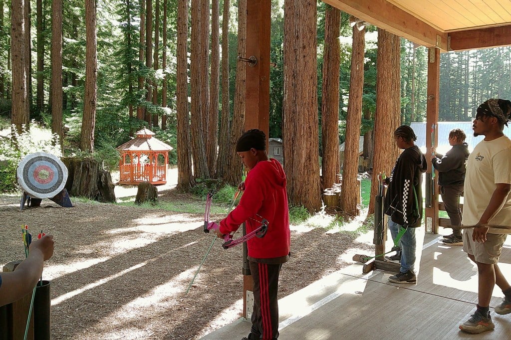 A young boy preps a bow with an arrow surrounded by wilderness.