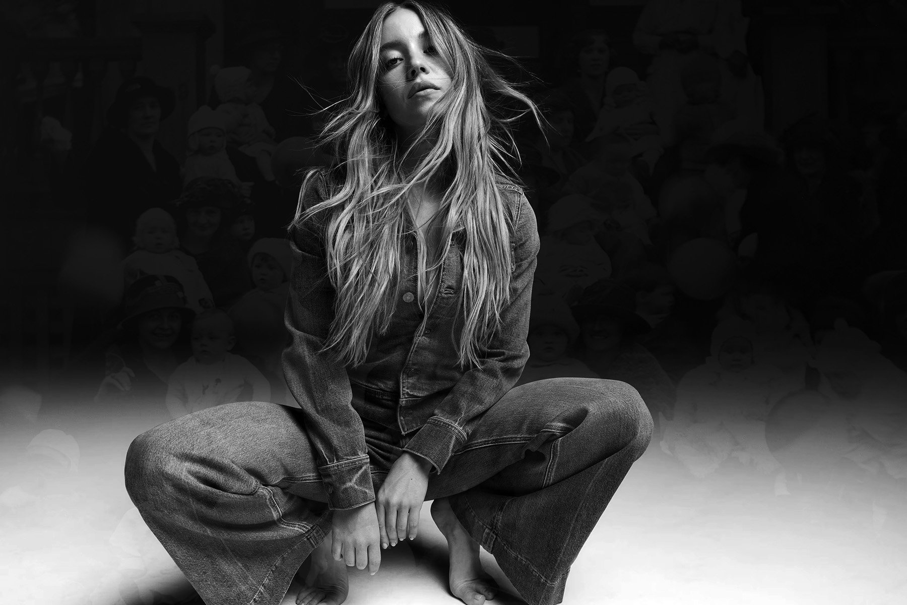 Sydney Sweeney, with long, loose hair squats barefoot in a wide-legged pose while wearing a denim jacket and flared jeans. She looks directly at the camera with a neutral expression. The background is a faded black-and-white photo of women and children.
