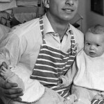 An archival image from the 1950s where a man is seen holding two babies and wearing an apron.