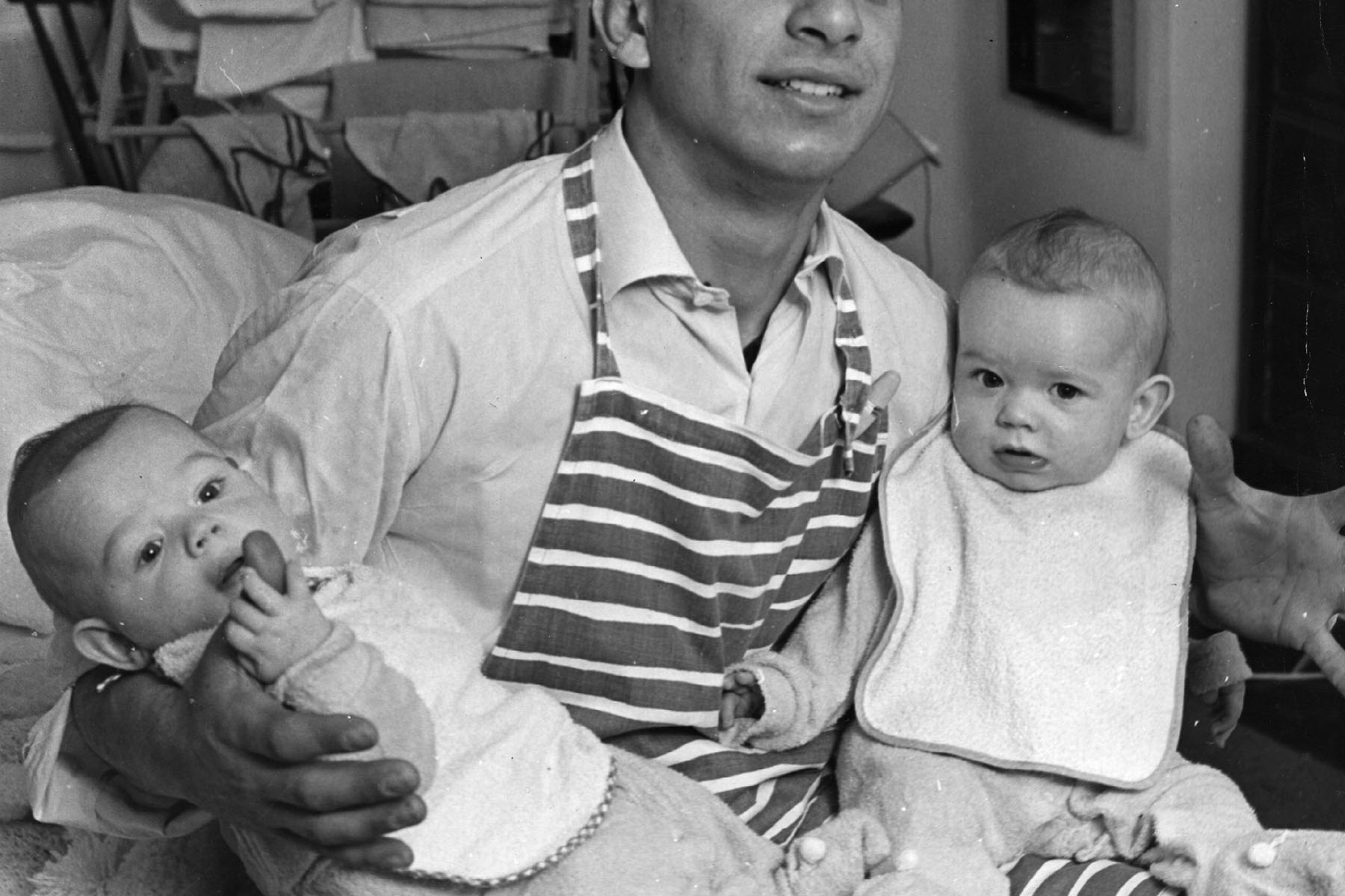 An archival image from the 1950s where a man is seen holding two babies and wearing an apron.
