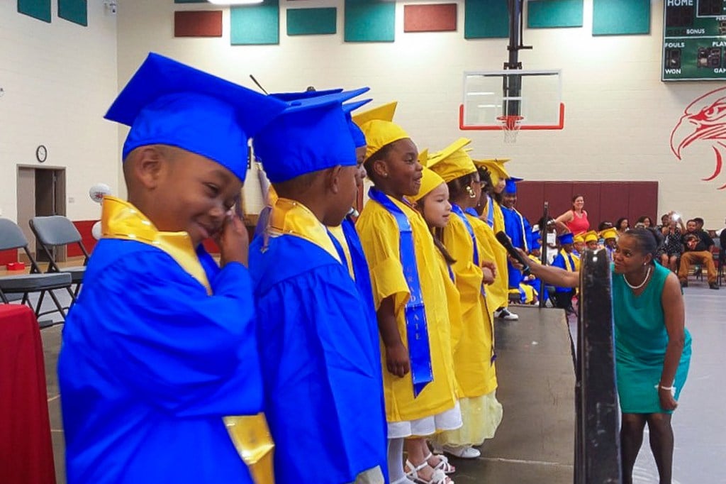 Several young children stand on a stage wearing graduation robes.