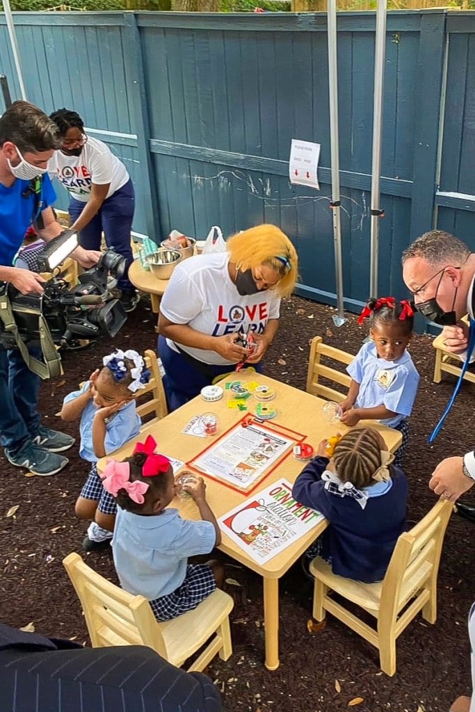 Young children sit at a table outside while caregivers in masks engage with them.